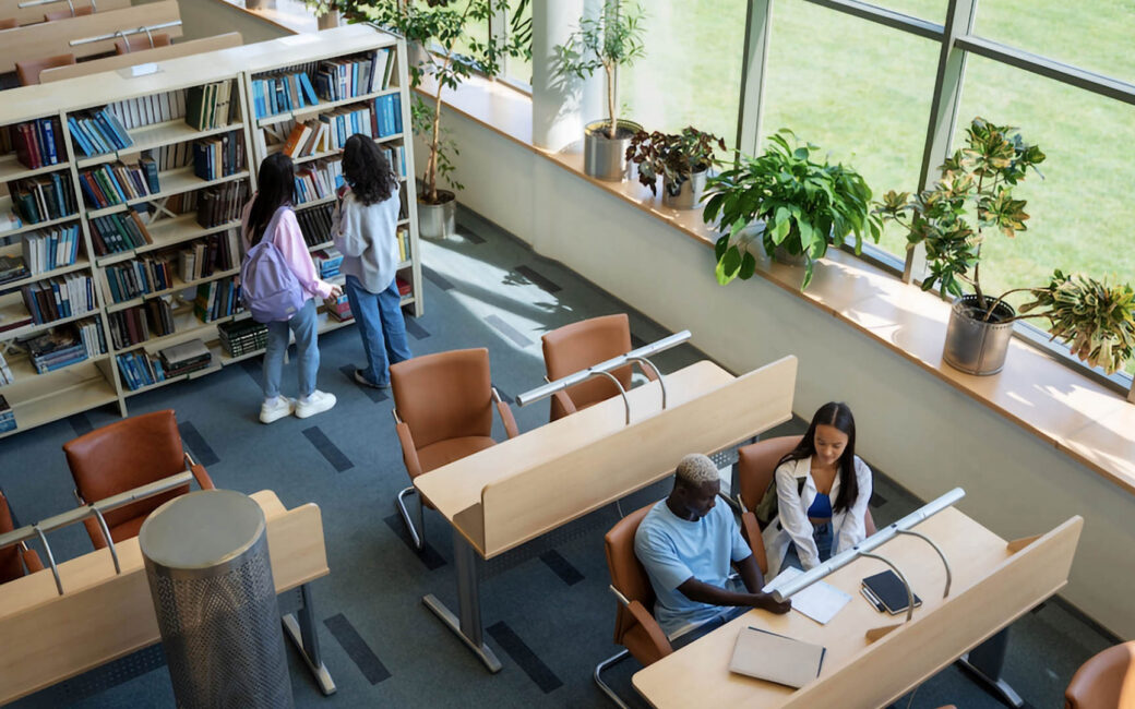 a school library with two students examining a bookshelf and two other students seated at a desk and reviewing notes