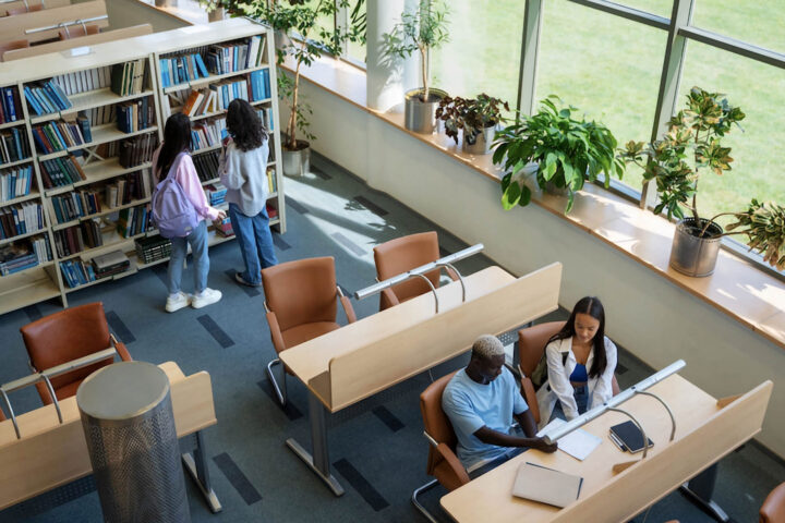 a school library with two students examining a bookshelf and two other students seated at a desk and reviewing notes